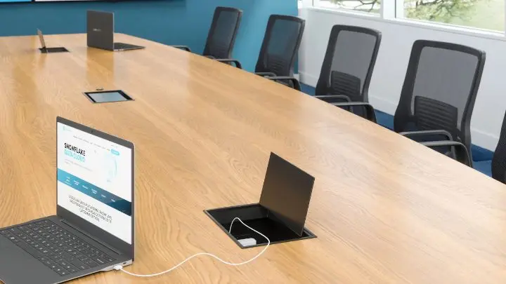 Close-up rendering of a light-oak conference table with a flip-up power and data box in the surface, a laptop, a tablet control panel and mesh task chairs along the far edge