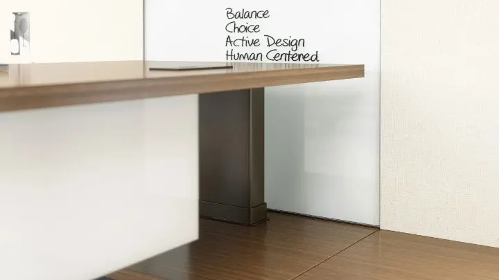 Close-up rendering of a standing-height wood worksurface on a metal panel leg, mounted against a whiteboard wall lettered with the words Balance, Choice, Active Design and Human Centered
