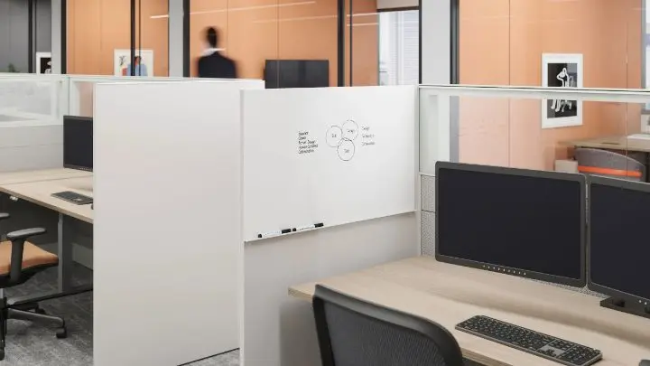 Close-up rendering of a panel-based workstation with a whiteboard panel and glass topper screen, a light-oak worksurface, dual monitors and a black mesh task chair, in an open-plan office