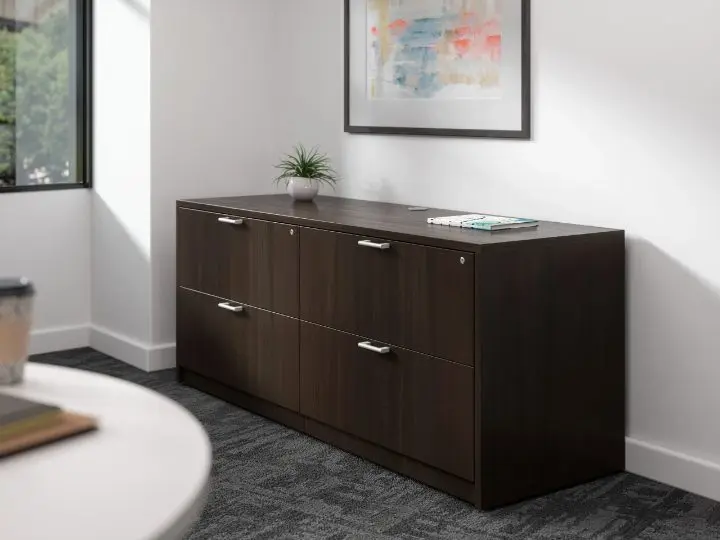 Close-up rendering of a dark espresso four-drawer lateral credenza with brushed metal pulls, set against a white wall below a framed print, with a round white table in the foreground