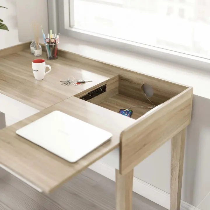 Close-up rendering of a light-oak desk with a sliding top panel that reveals a cable and device trough, shown with a laptop mat, a pen cup and a mug at a sunlit window
