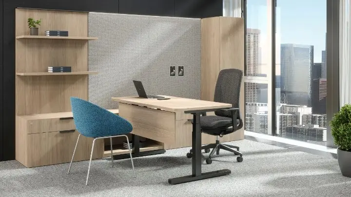 Private office in a pre-designed setting with a height-adjustable light-oak desk, a low credenza and wall-mounted shelving, an acoustic fabric panel, a dark mesh task chair and a teal guest chair, with a city view through floor-to-ceiling glass