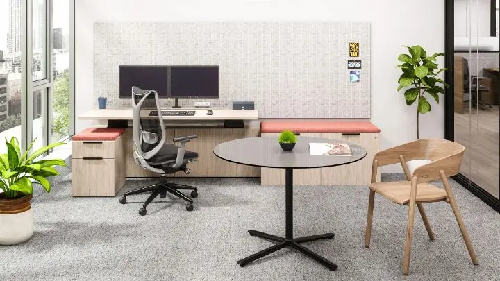 Workstation in a pre-designed setting with a light-oak height-adjustable desk, dual monitors on an arm, a tackable fabric wall panel, a red upholstered window bench, a mobile pedestal and a round meeting table with a wood-frame guest chair
