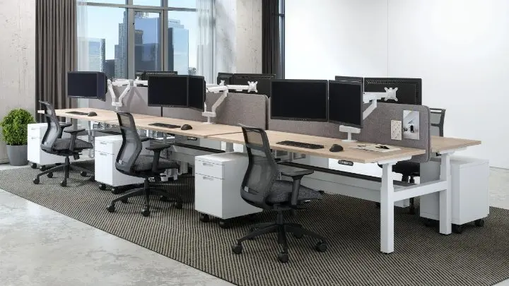 Six-person bench workstation run in a pre-designed setting with light-oak tops on a white frame, grey acoustic divider screens, dual monitor arms, white mobile pedestals and black mesh task chairs, with a city view