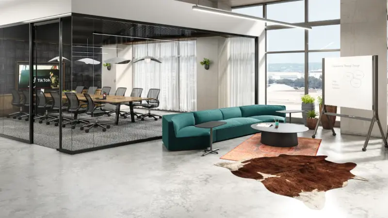 Glass-walled conference room rendering with a teal modular sofa and mobile whiteboard, Anomatics