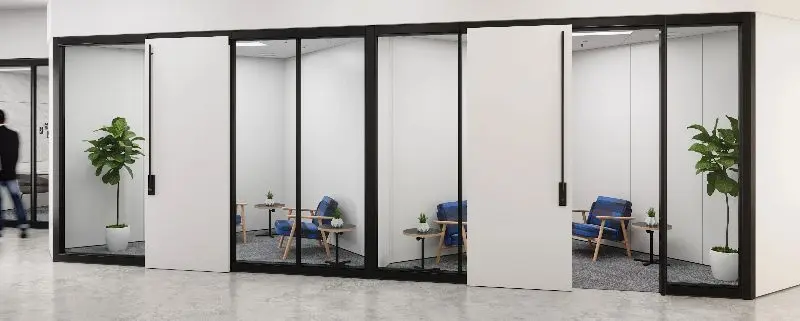 Divider wall system rendering with black-framed glass fronts and sliding solid panels on a row of two-person phone rooms with blue lounge chairs, side tables and planters